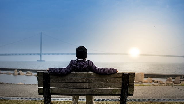 Man looking at the Sun setting below a bridge while he sits on a bench at a coast, resting his feet on grass, wearing a jacket and a cap, because apparently it is really cold at the spot of the picture and it seems like this picture was not taken in India&nbsp;