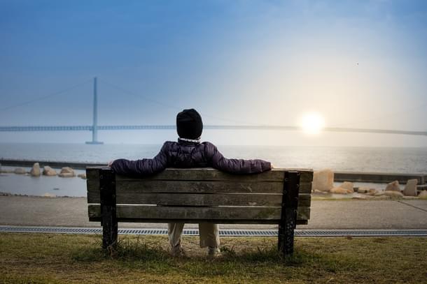 Man looking at the Sun setting below a bridge while he sits on a bench at a coast, resting his feet on grass, wearing a jacket and a cap, because apparently it is really cold at the spot of the picture and it seems like this picture was not taken in India 